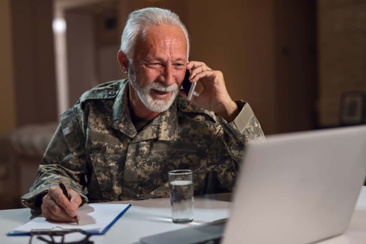 Smiling veteran communicating on mobile phone while doing paperwork and using laptop in the office.