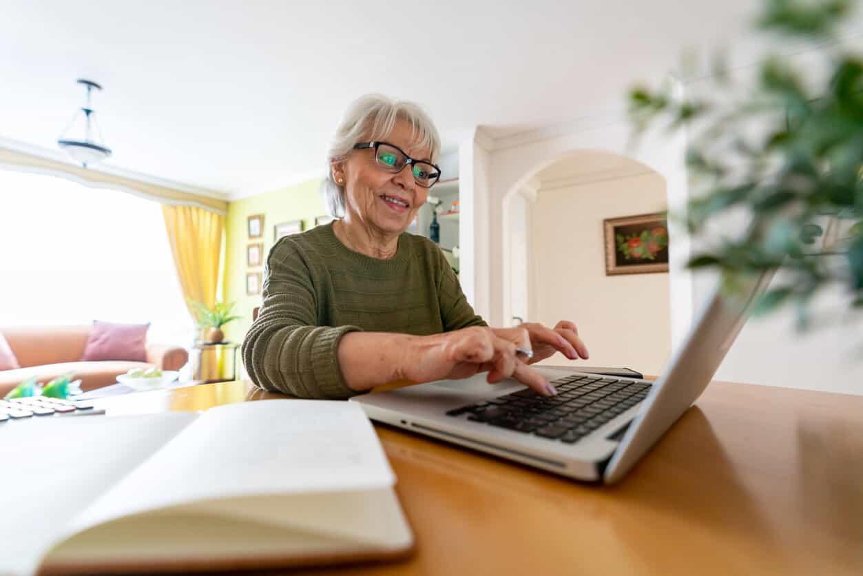 Senior Woman Using Laptop for Obituaries Elderly woman browsing obituaries on her laptop at home, smiling and engaged in online research.