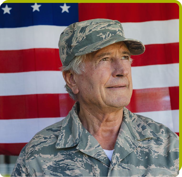 Veteran in Military Uniform Senior military veteran in camouflage uniform standing in front of an American flag, symbolizing service and patriotism.
